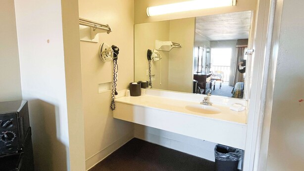 A bathroom with a white sink, a mirror, a hairdryer, and a trash can. - Motel 6 Bracey, VA – I-85 (Bracey)
