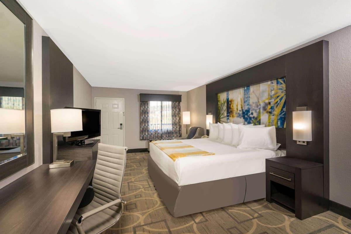 A hotel room with a bed, a desk with a TV, a chair, a desk lamp, a bedside table, and a painting on the wall.
