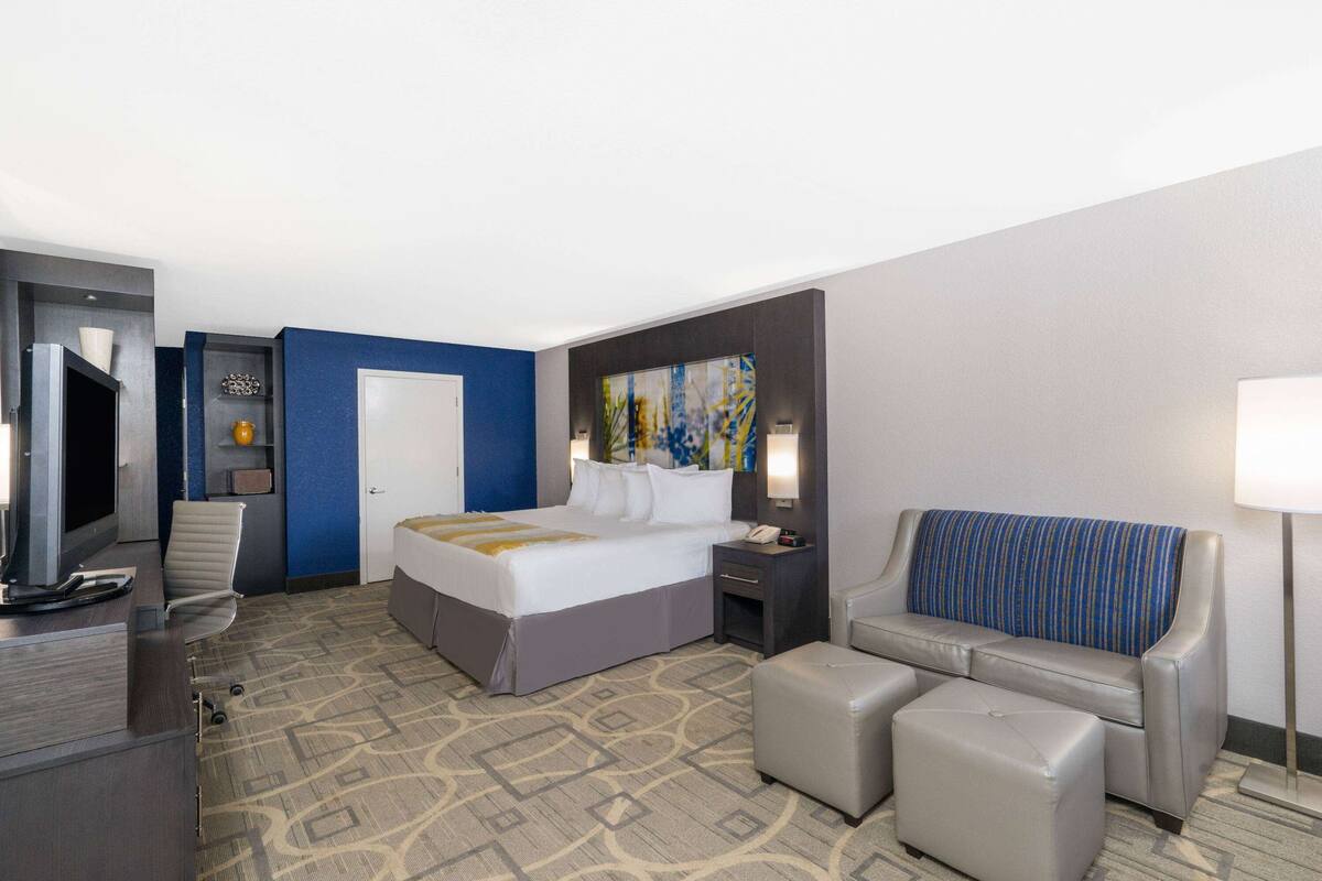 A hotel room with a bed, a desk with a computer, a chair, a sofa, and a painting on the wall.