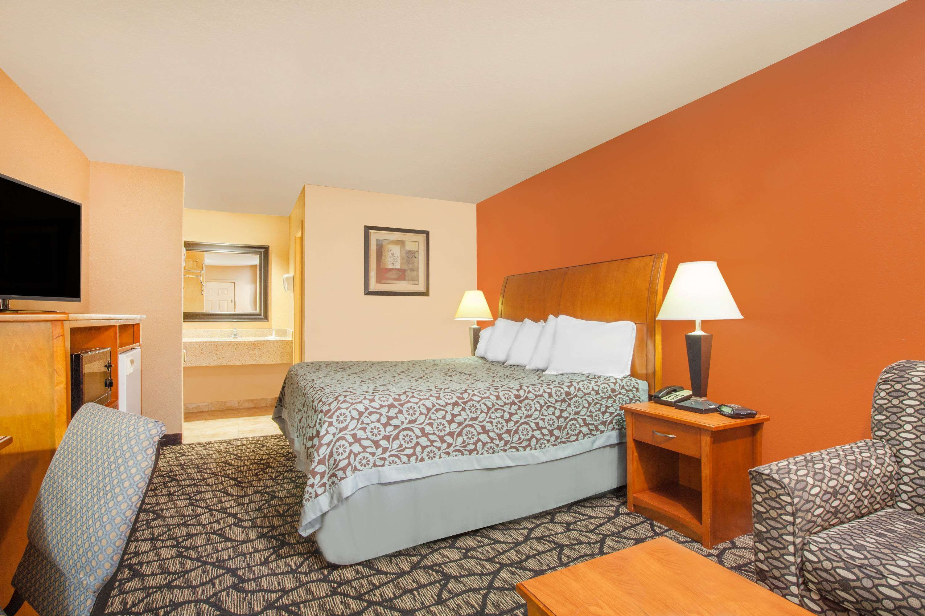 Une chambre d’hôtel comprenant un lit, un bureau avec un téléviseur à écran plat, une chaise, une table de chevet avec une lampe, et une salle de bain visible à travers une porte ouverte.