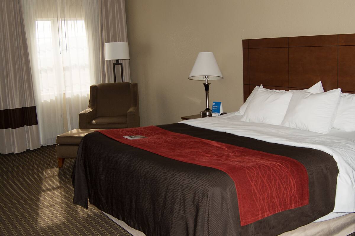 Une chambre d’hôtel avec un grand lit, une chaise, une table de chevet, une lampe et une fenêtre avec des rideaux.