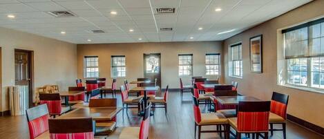 A spacious dining area with multiple tables and chairs, large windows, and a high ceiling with recessed lighting.
