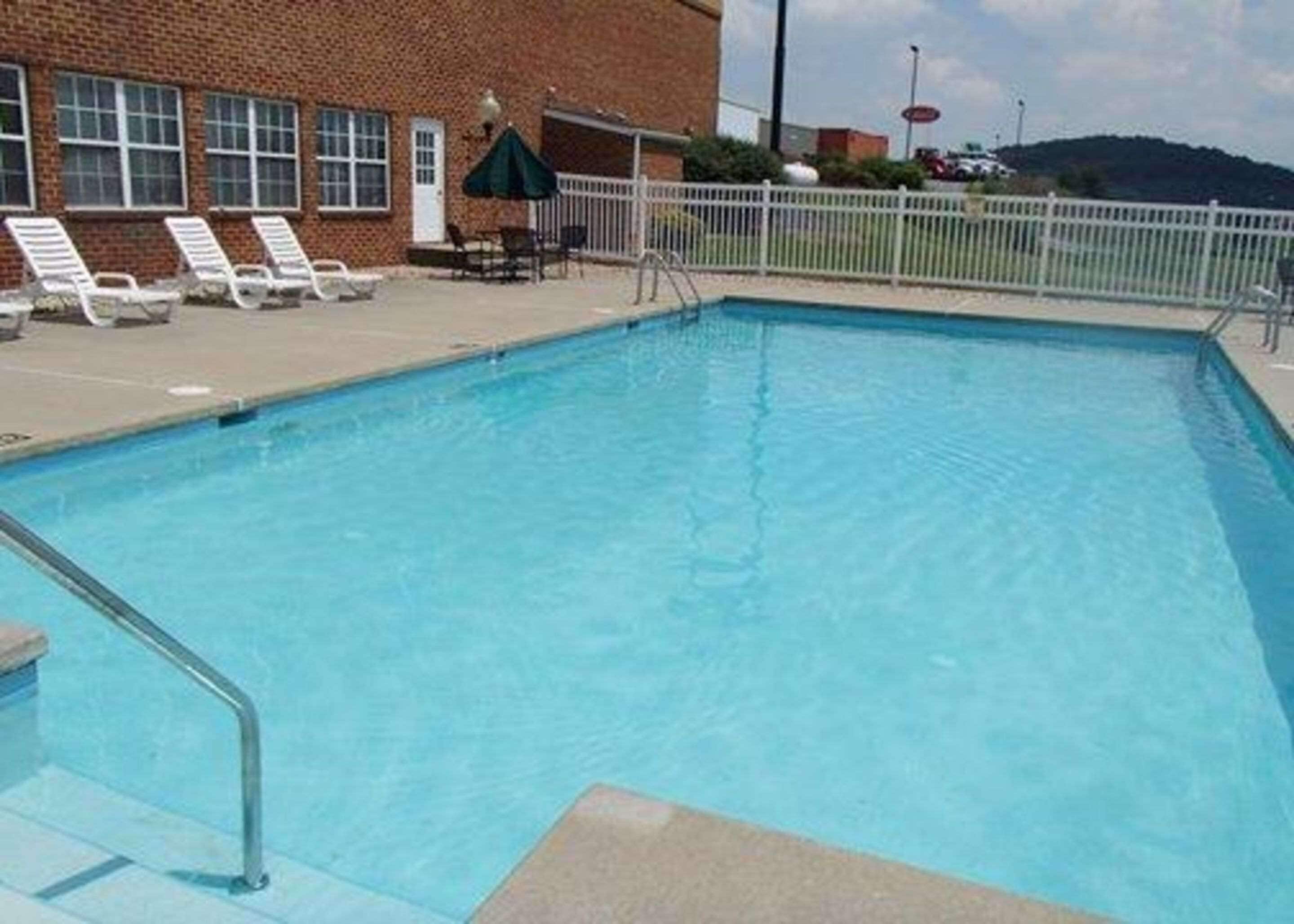 A rectangular swimming pool with a metal ladder, surrounded by a white fence. There are lounge chairs and a table with a green umbrella.