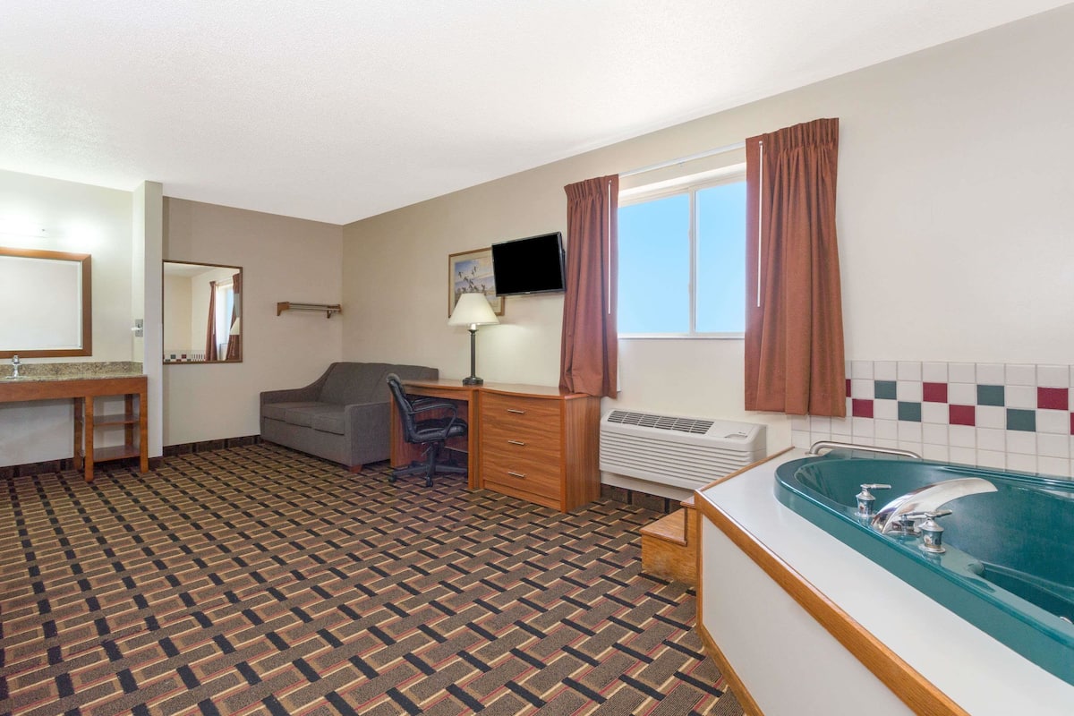 A hotel room with a patterned carpet, a desk with a chair, a television, a large window with curtains, and a bathtub.