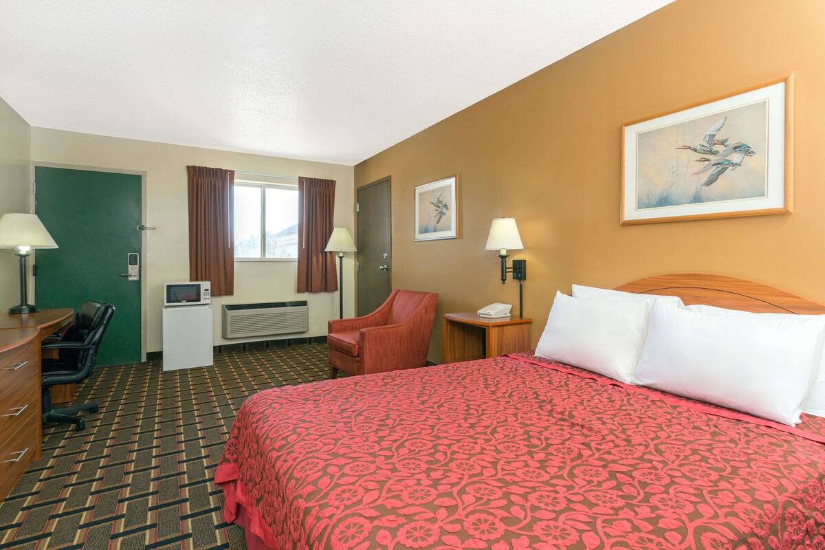 A hotel room with a large bed, a desk with a chair, a TV, a telephone, and a window with curtains.