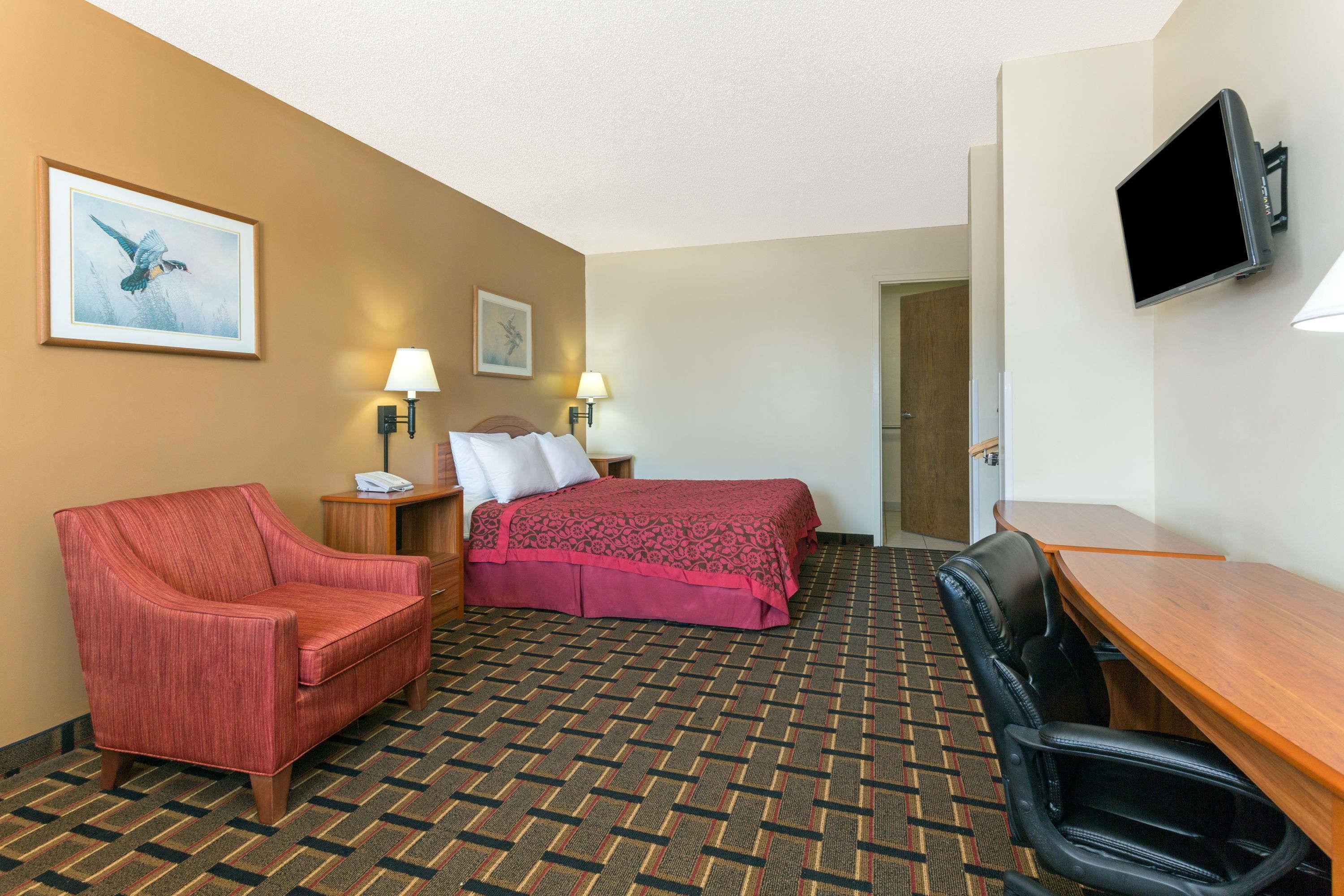 Habitación de hotel con cama, escritorio, silla, sillón rojo, dos lámparas de pared y televisión.