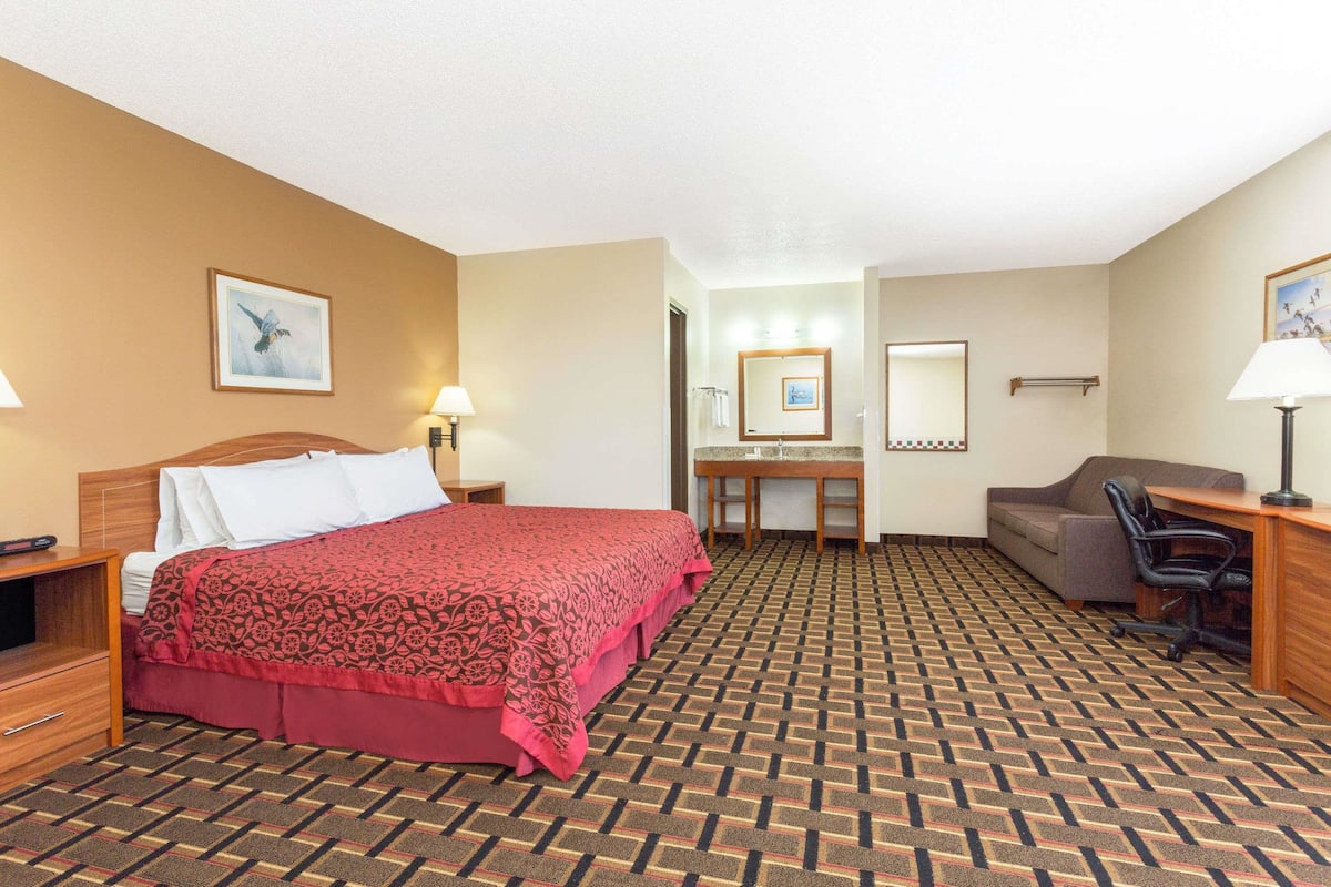 A hotel room with a large bed, a desk, a chair, a sofa, and a bathroom visible through an open door.