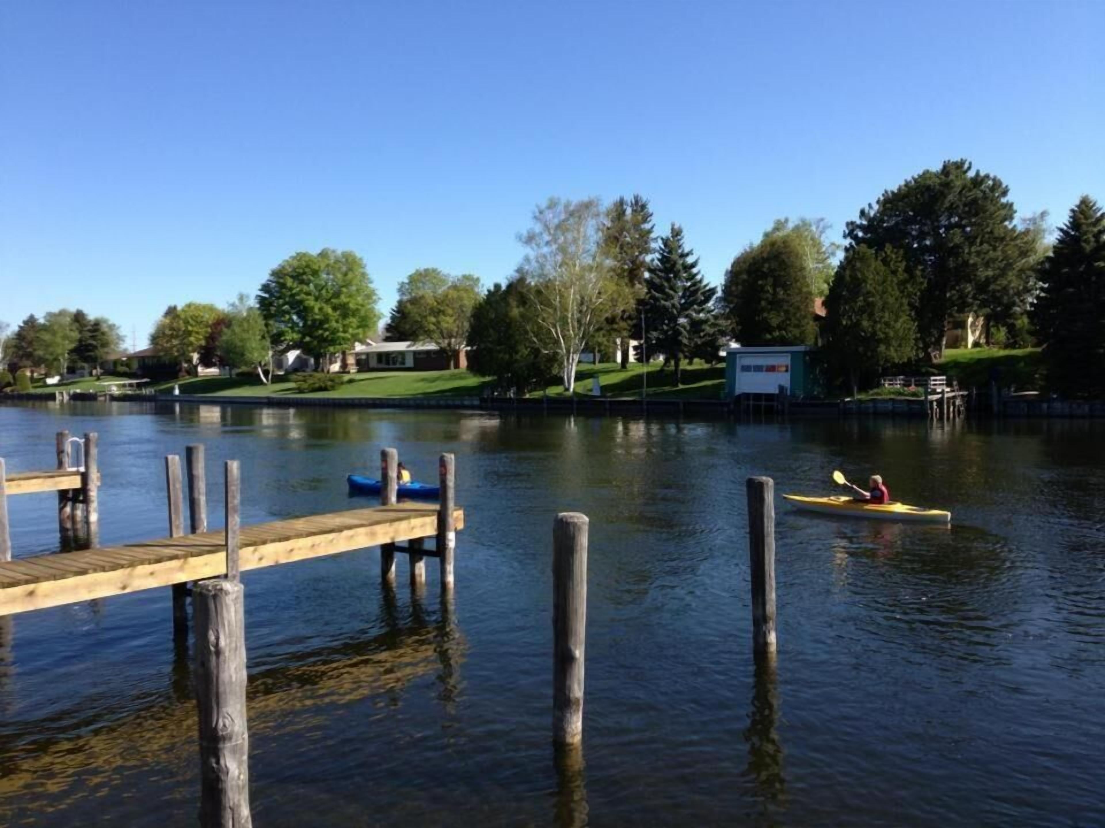 Un lago tranquilo con un muelle de madera que se adentra en el agua. Hay kayaks flotando y un pequeño edificio en la orilla.