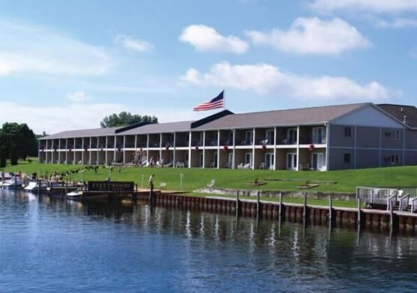 A motel with a large American flag, a dock with boats, and a grassy area in front. - Fleetwood Inn & Suites (Cheboygan)