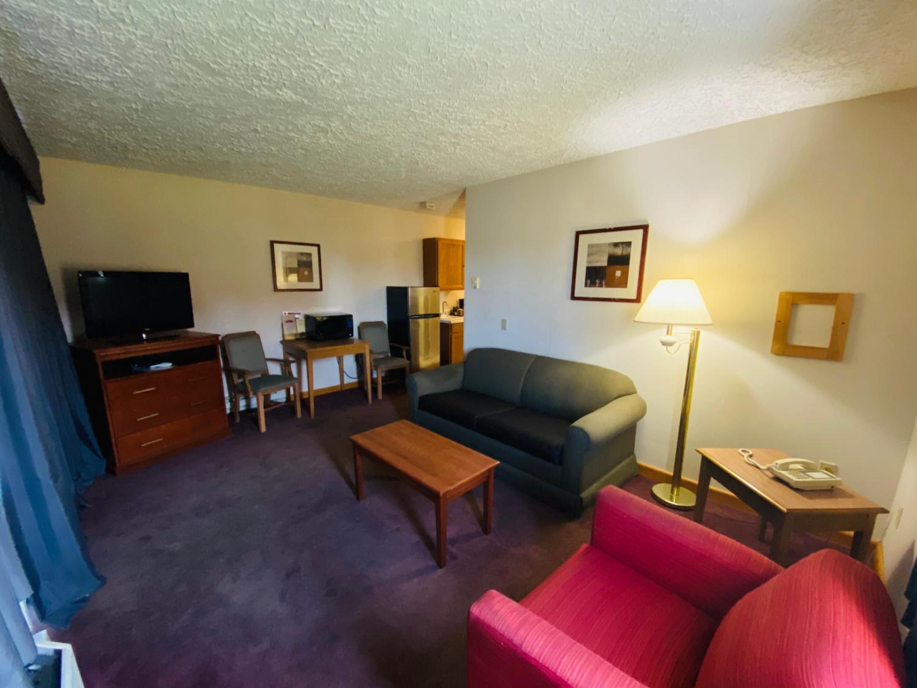 A compact living room with a sofa, a red armchair, a wooden coffee table, a TV on a cabinet, a desk with a chair, a refrigerator, and a lamp.