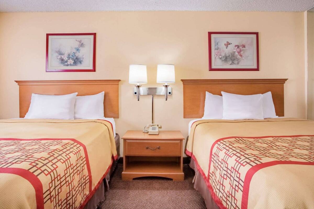 A hotel room with two beds, a nightstand with a lamp, and two framed floral artworks on the walls.
