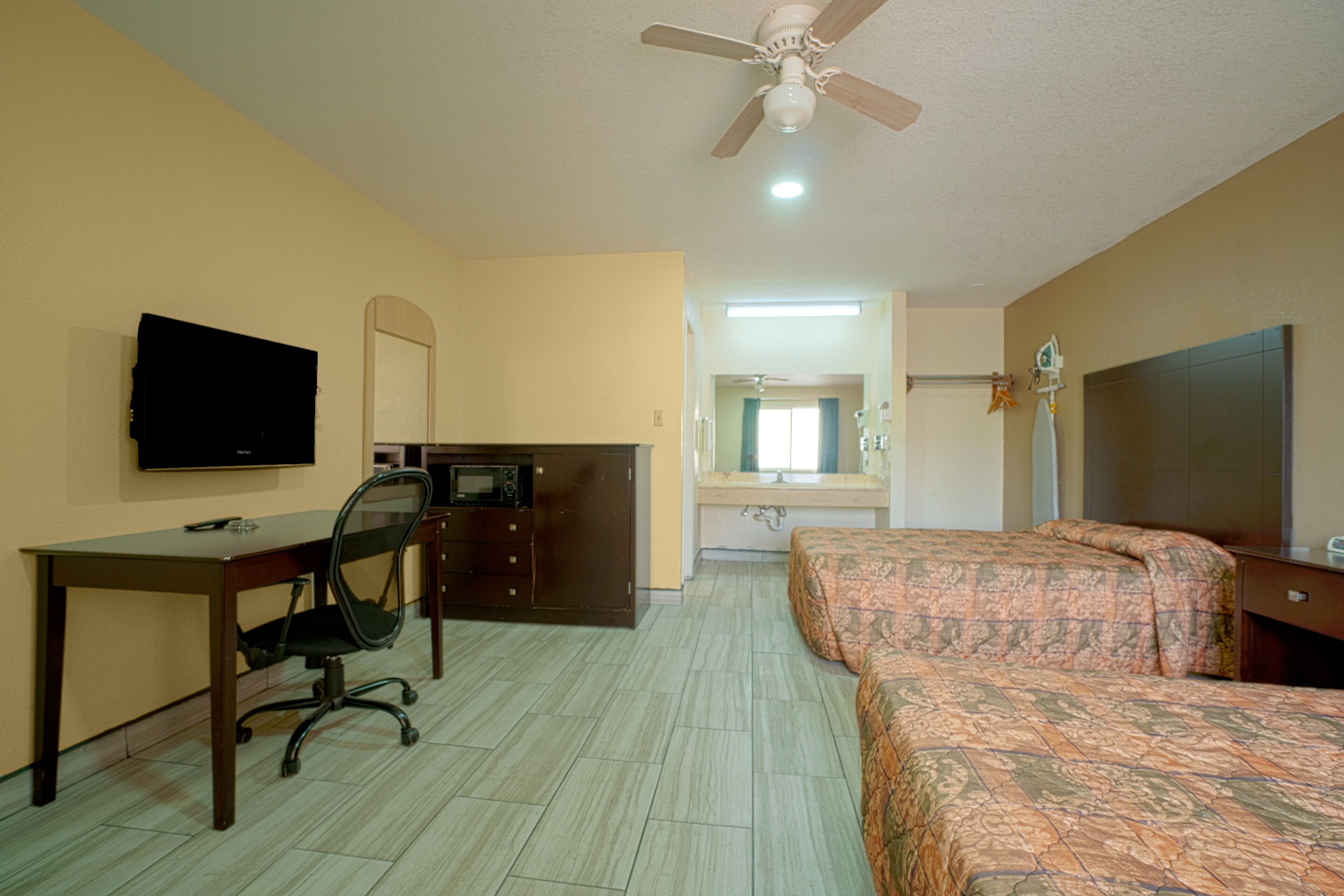 A hotel room with a desk, chair, bed, ceiling fan, and a television.