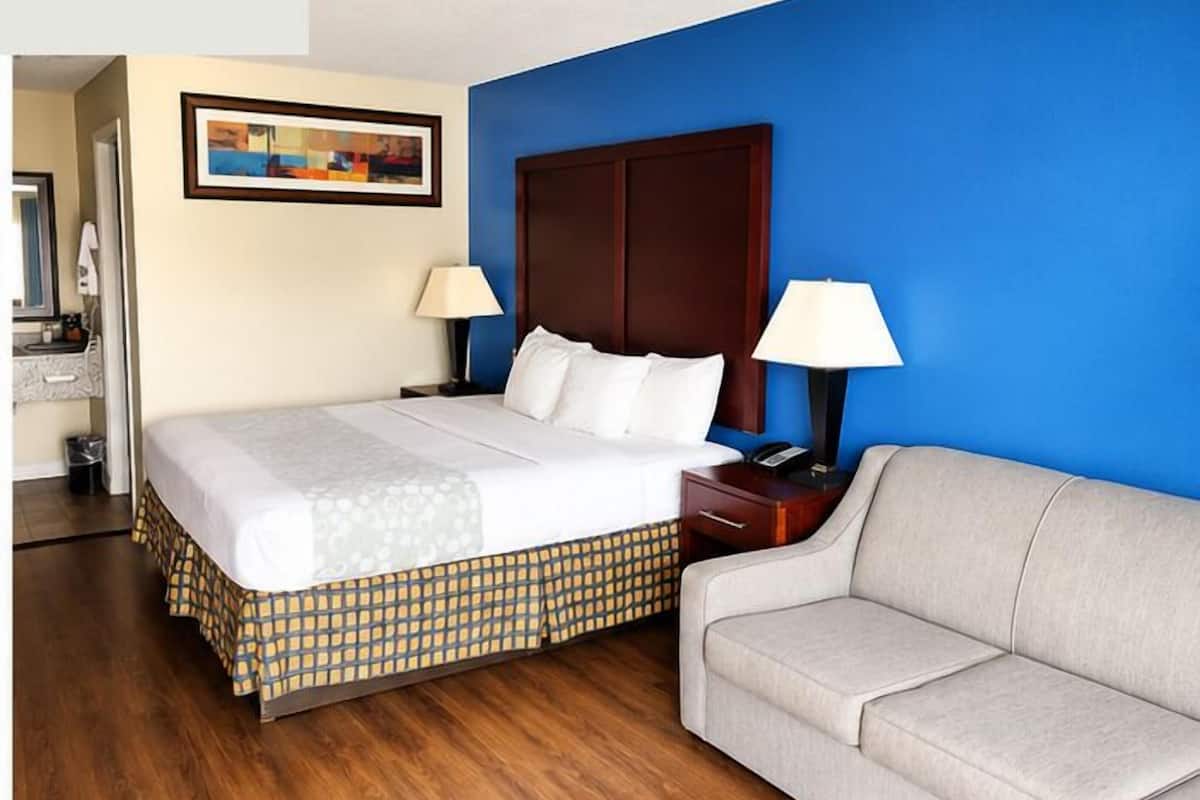 A hotel room with a bed, a sofa, a nightstand, a lamp, a painting, and a wooden floor.
