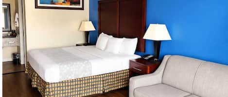 Habitación de hotel con cama, sofá, mesita de noche, lámpara, un cuadro y suelo de madera.