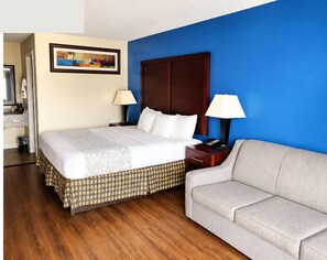 Habitación de hotel con cama, sofá, mesita de noche, lámpara, un cuadro y suelo de madera.