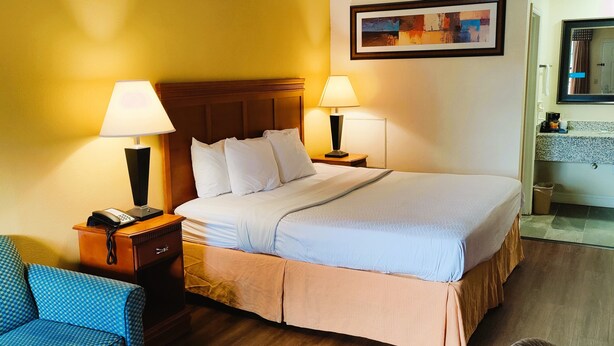 Una habitación de hotel con una cama grande, dos lámparas de noche, una mesita de noche de madera, un sillón azul estampado y una pequeña mesa de madera.