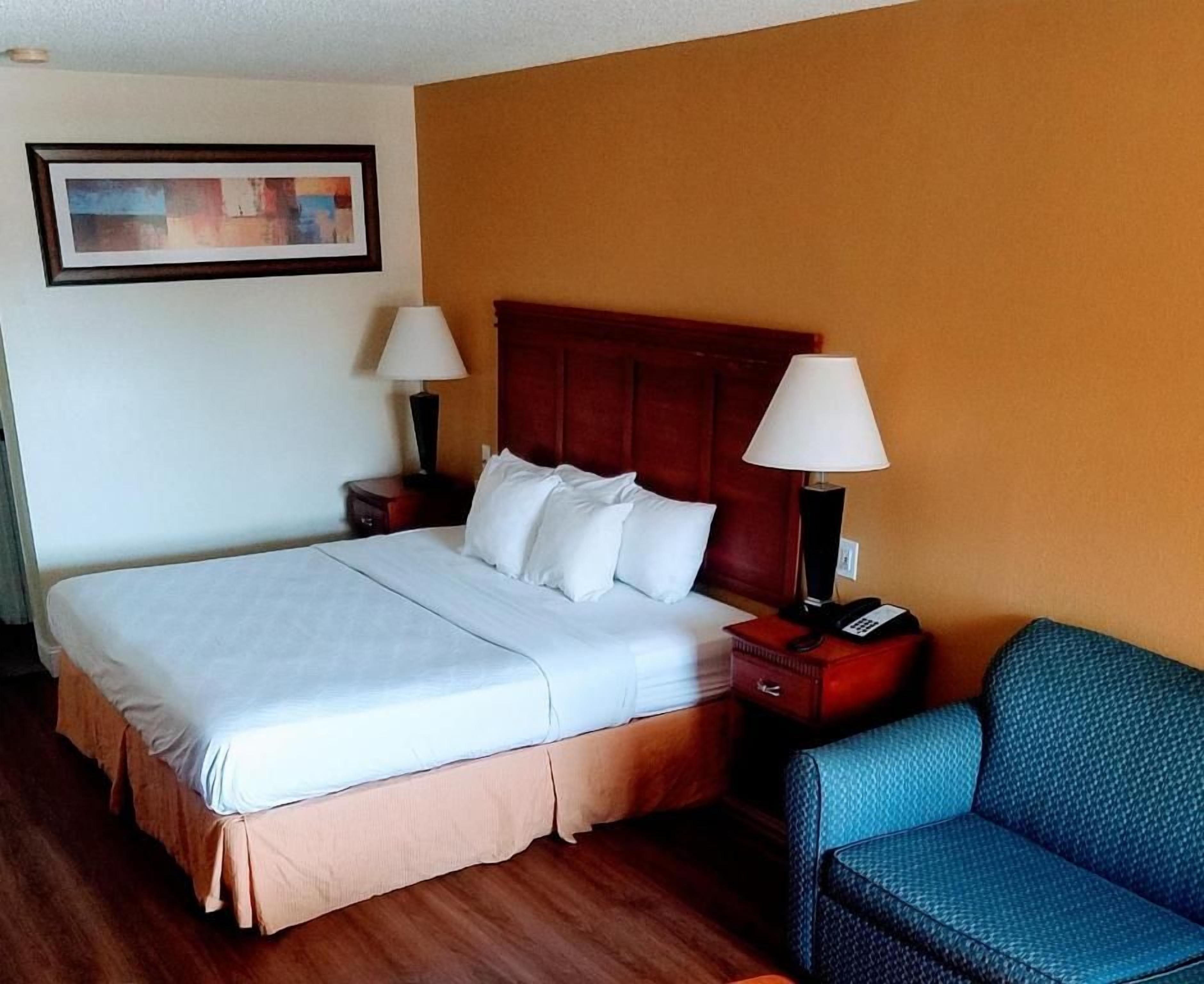 A hotel room with a large bed, two bedside tables with lamps, a blue armchair, and a framed picture on the wall.