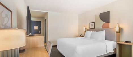 Habitación de hotel con una cama grande, un escritorio con lámpara, un televisor y una puerta que da a otra habitación.
