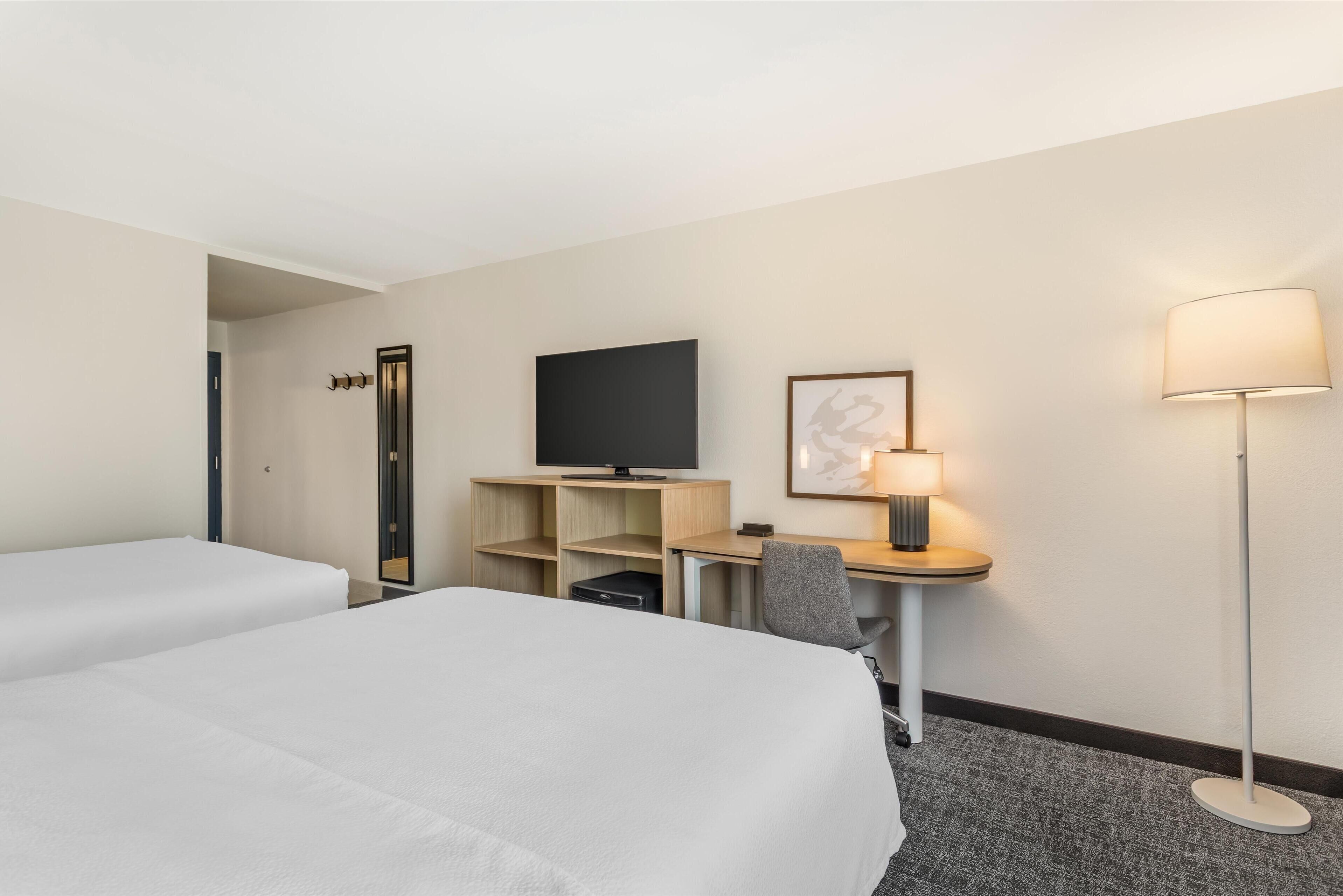 Ein Hotelzimmer mit zwei Betten, einem Schreibtisch mit Computer, einem Fernseher, einer Lampe und einer Tür.
