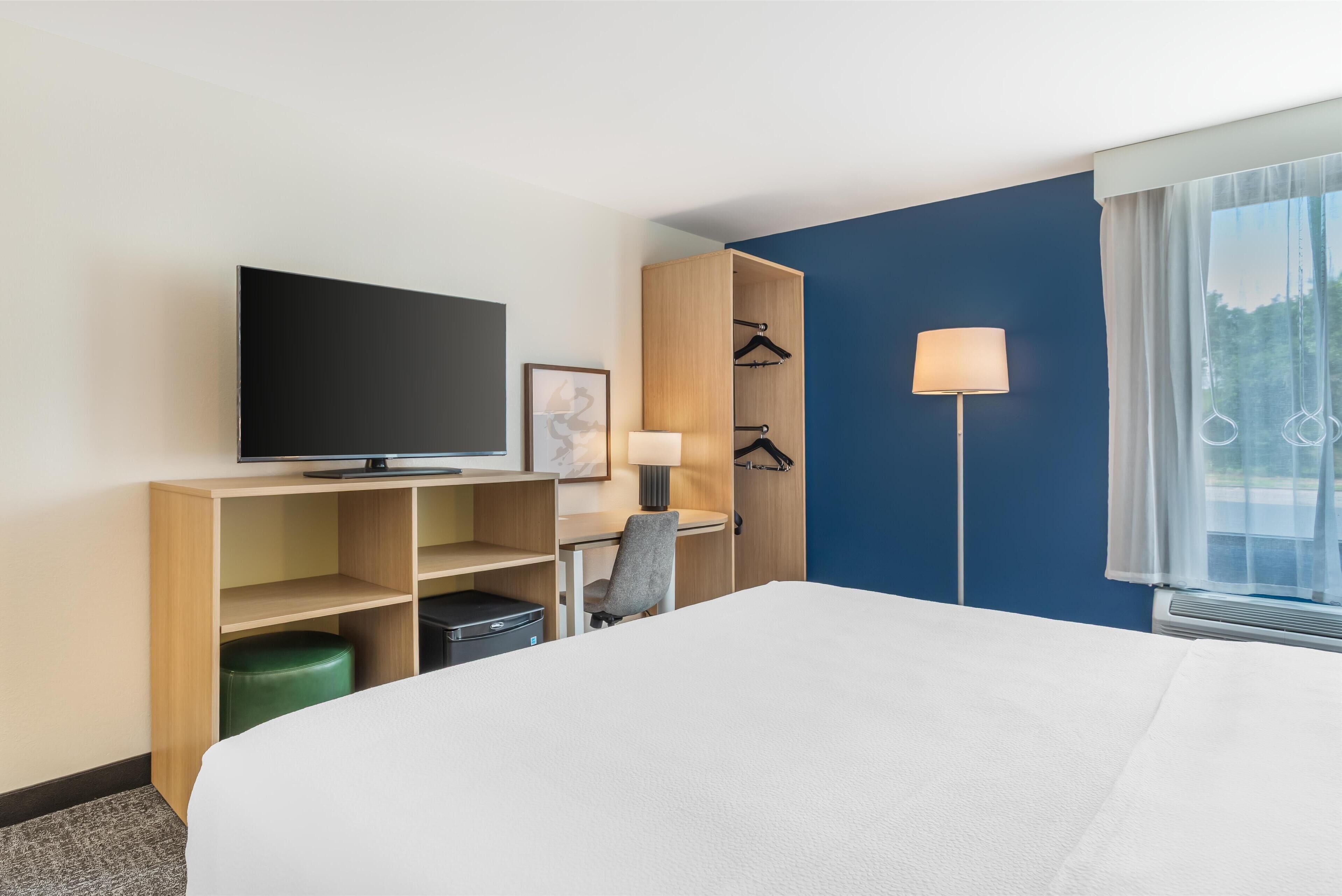 Hotellihuone, jossa on sänky, televisio, työpöytä, tuoli, lamppu, vaatekaappi ja ikkuna, jossa on verhot.