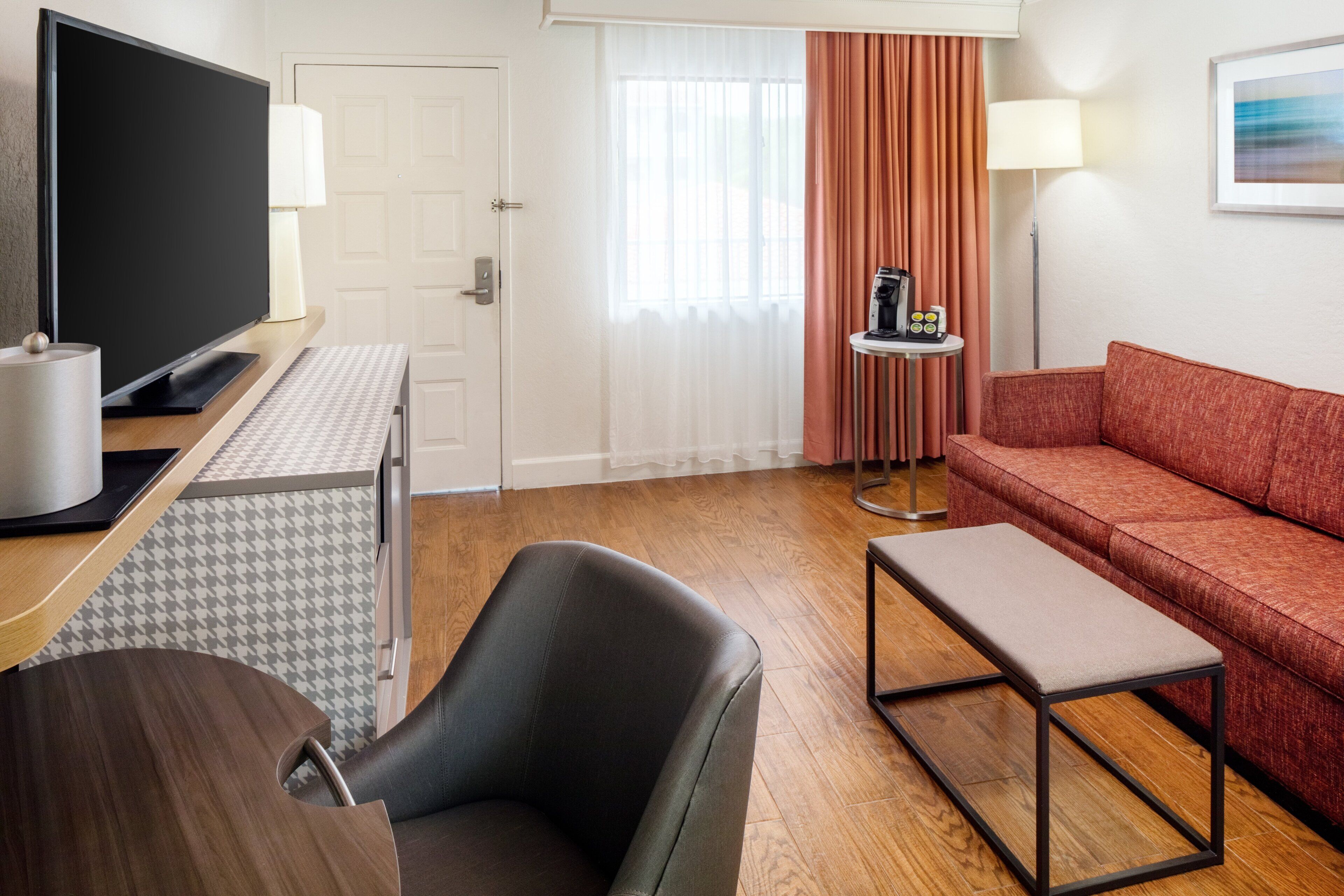 A hotel room with a flat-screen TV, a sofa, a chair, a small table, and a coffee machine.