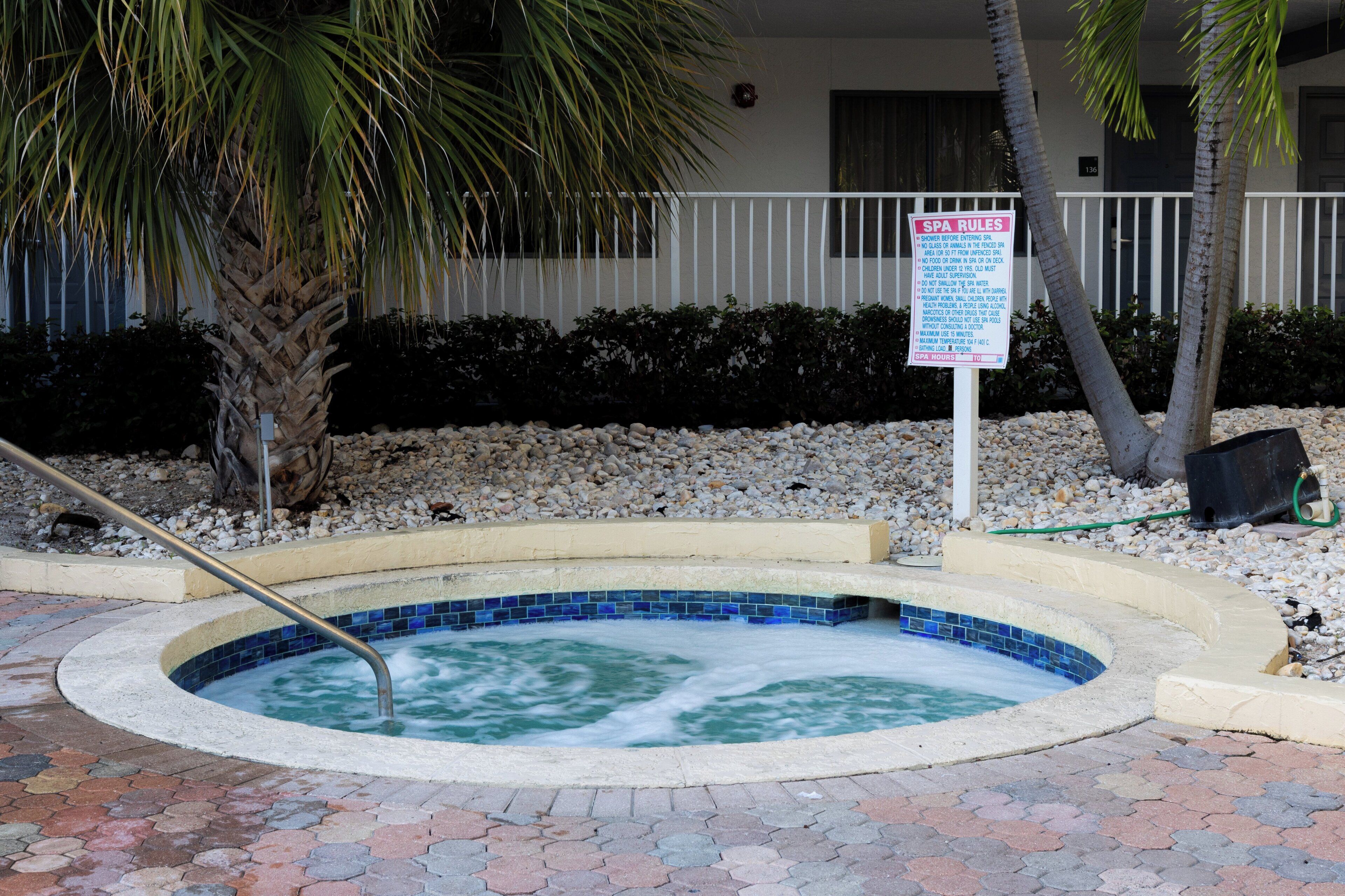 A hot tub with a sign indicating spa rules, surrounded by a pebble bed and palm trees.