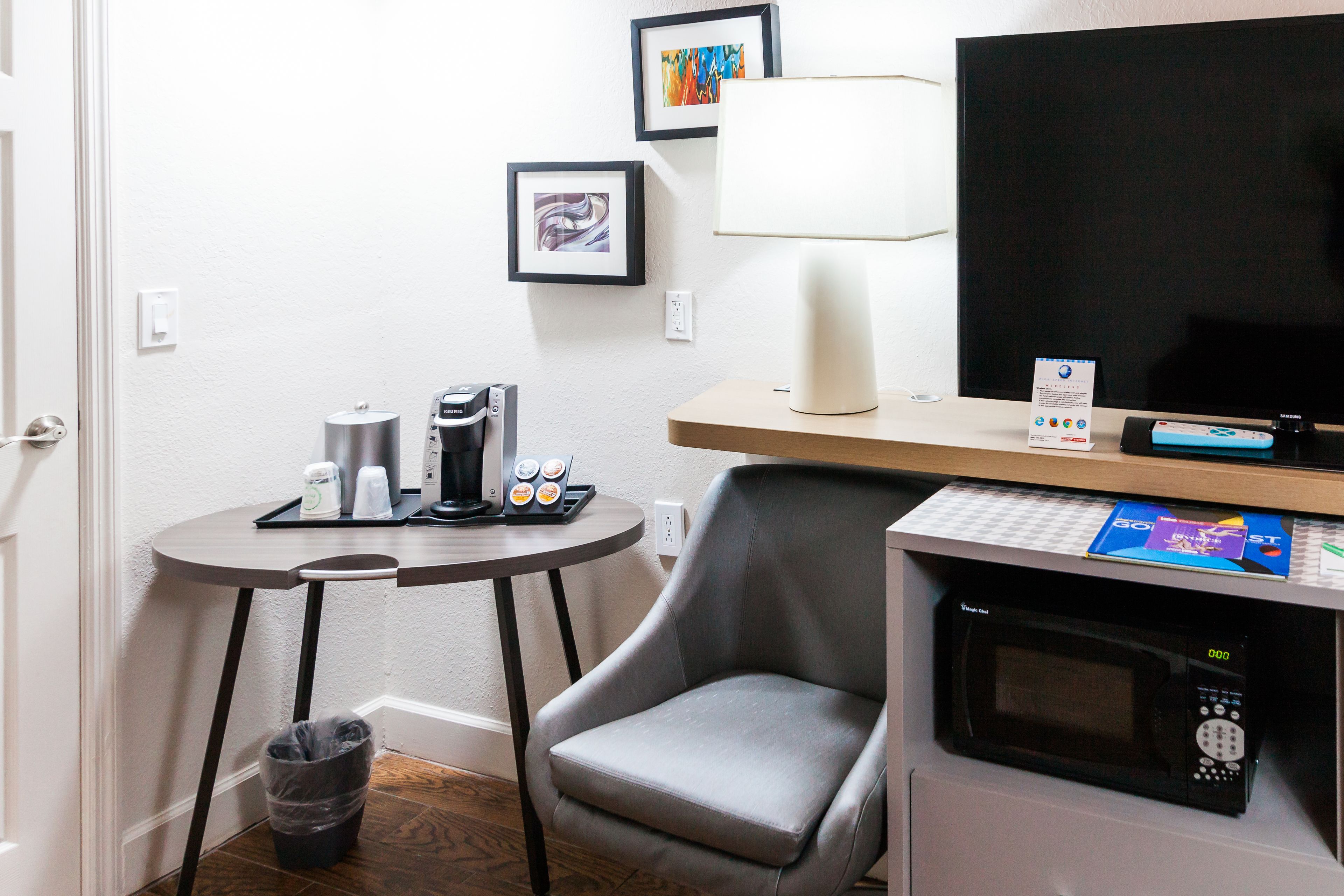 A compact hotel room with a small table, a microwave, a chair, a trash bin, a TV, and a lamp.