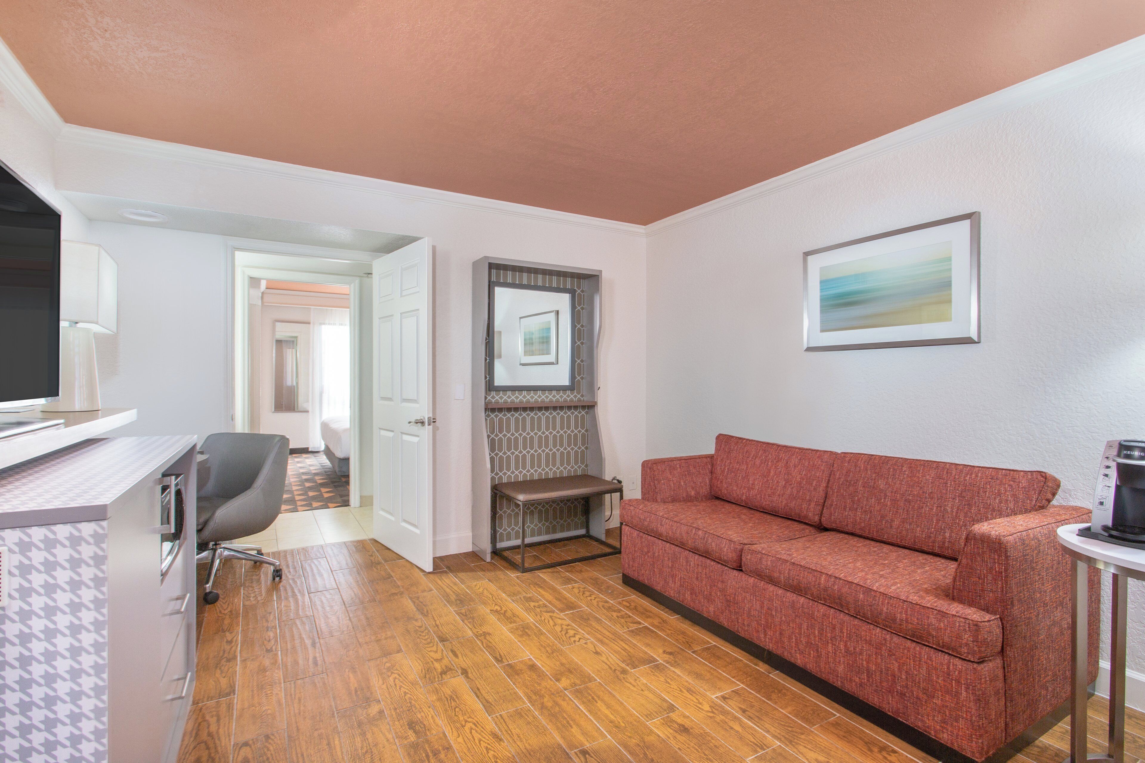Une chambre d’hôtel avec un plancher en bois, un canapé rouge, un bureau avec un ordinateur, une machine à café et un tableau encadré au mur.