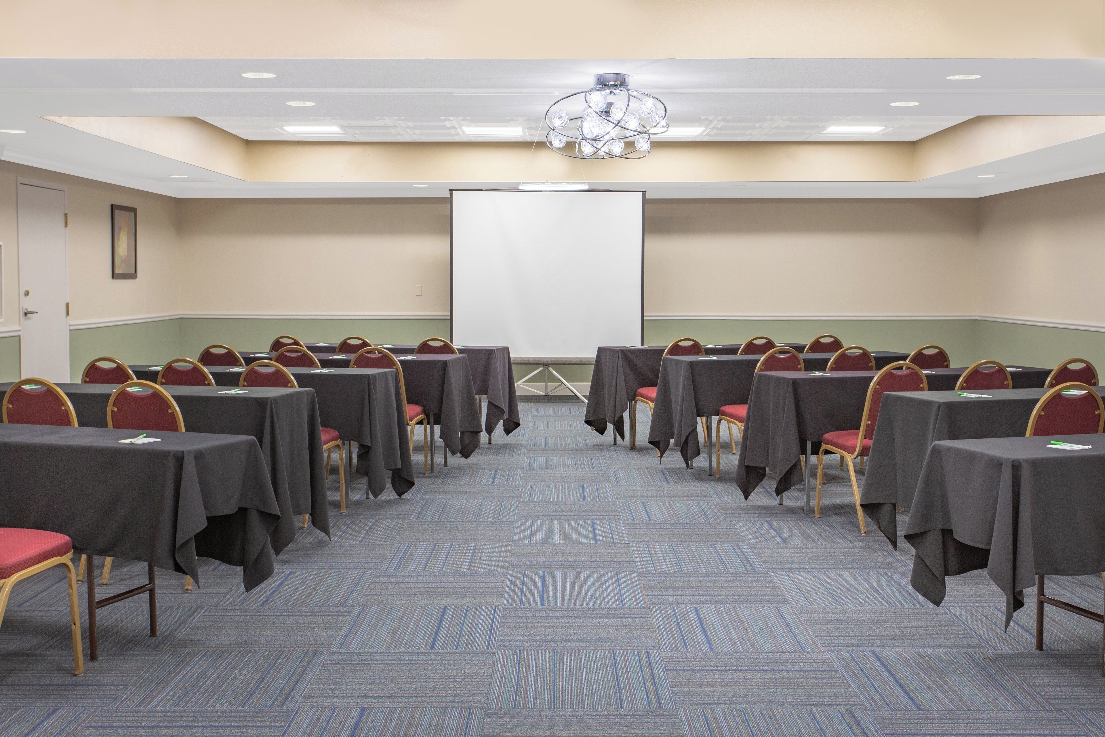 Un salón de conferencias con filas de mesas y sillas, una pantalla de proyector y una araña.