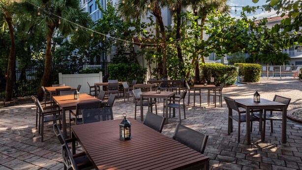 An outdoor patio area with multiple wooden tables and chairs, surrounded by lush greenery and palm trees. - Holiday Inn & Suites Boca Raton - North by IHG (Boca Raton)