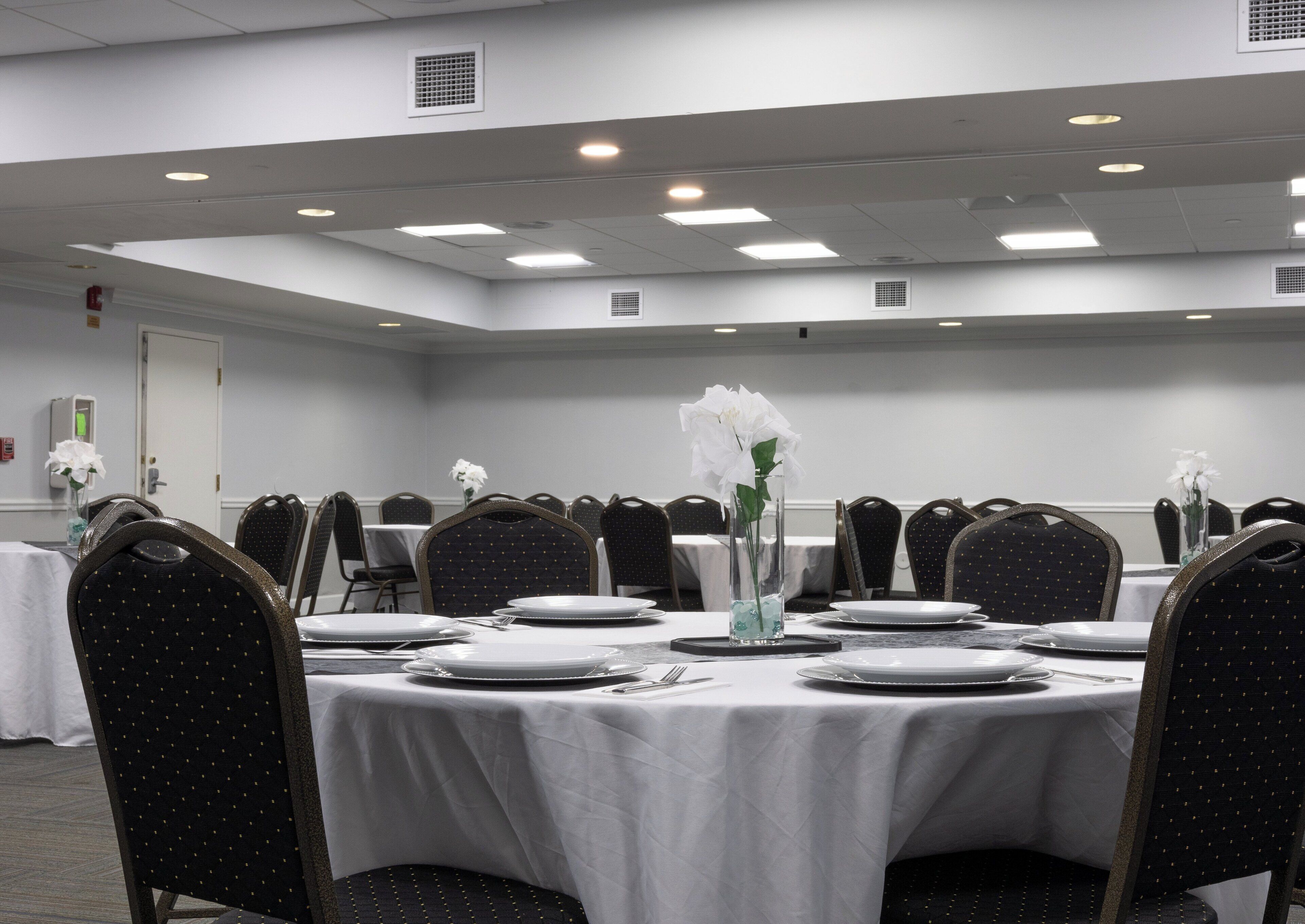 Un salón de conferencias con mesas redondas, sillas negras, manteles blancos y flores blancas en jarrones.