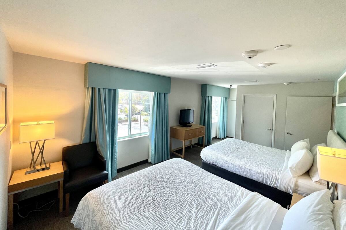Ein Hotelzimmer mit zwei Betten, einem Schreibtisch mit Lampe, einem Sessel, einem Fernseher und einem Kleiderschrank.