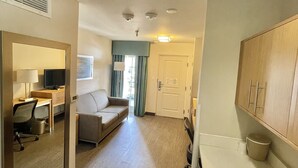 A hotel room with a kitchenette, a sofa, a desk with a computer, and a bathroom with a toilet and sink.