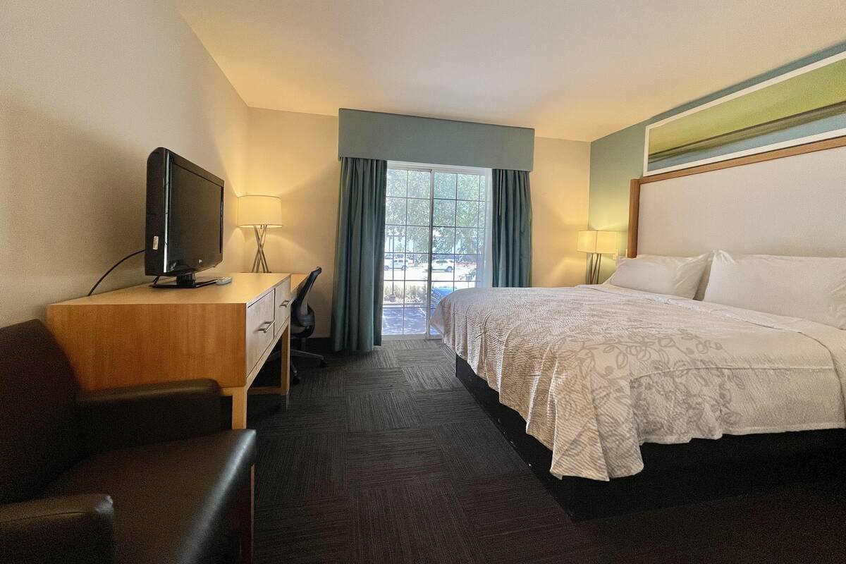 Ein Hotelzimmer mit einem Bett, einem Schreibtisch mit Fernseher, einem Sessel und einem Fenster mit Vorhängen.