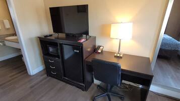 A hotel room with a desk, a TV, a lamp, a chair, a bed, and a bathroom with a sink and towel rack.