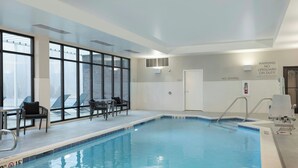 Ein Hallenbad mit deutlicher Sicherheitsschilderung, einem sonnendurchfluteten Sitzbereich und Blick durch große Fenster.