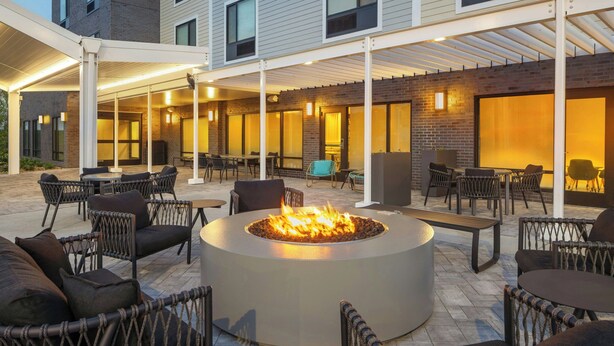 Ein Terrassenbereich im Freien mit einer runden Feuerstelle, umgeben von Korbstühlen und Tischen.