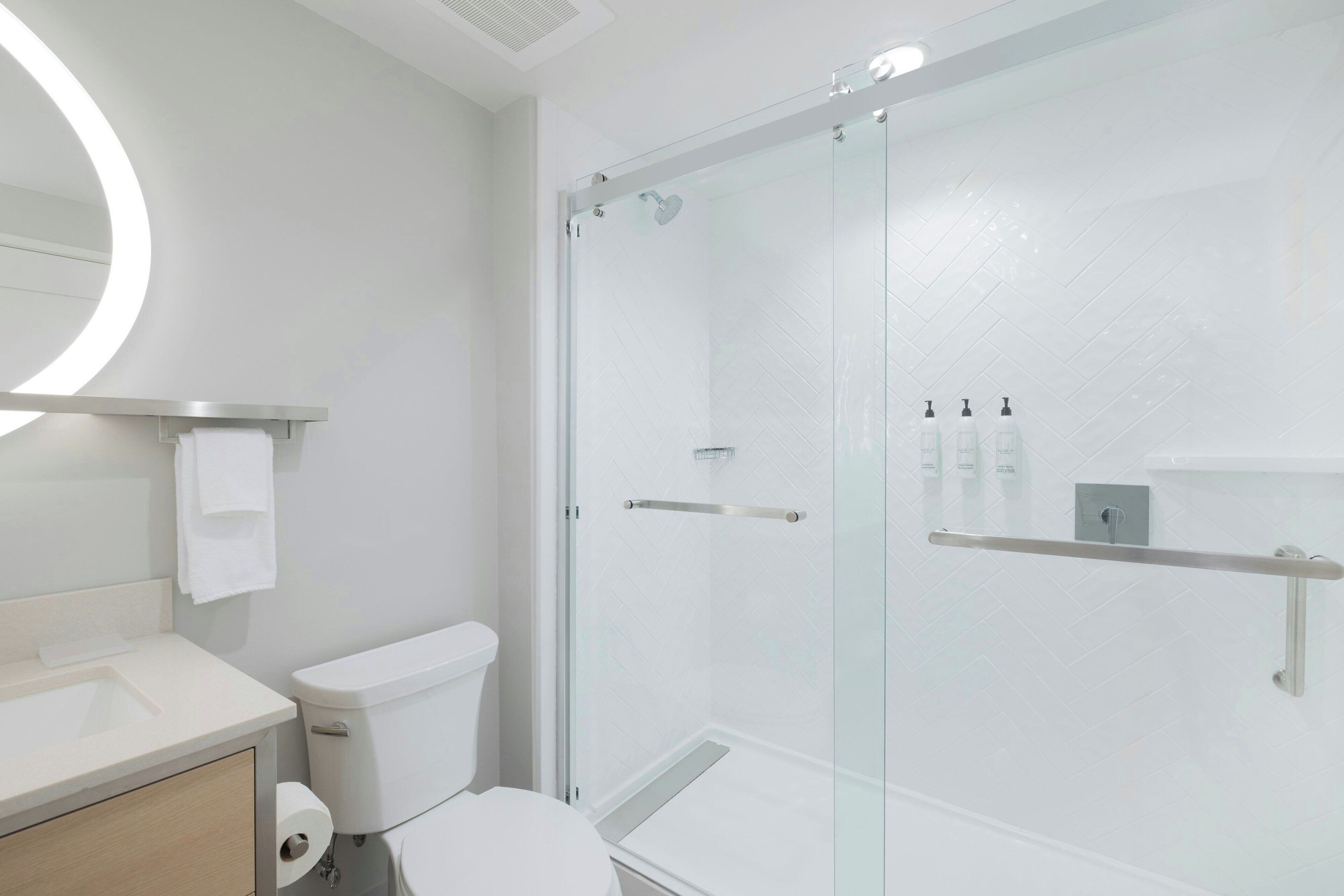 Un baño moderno con ducha de vidrio, inodoro, lavamanos y espejo con luz circular.