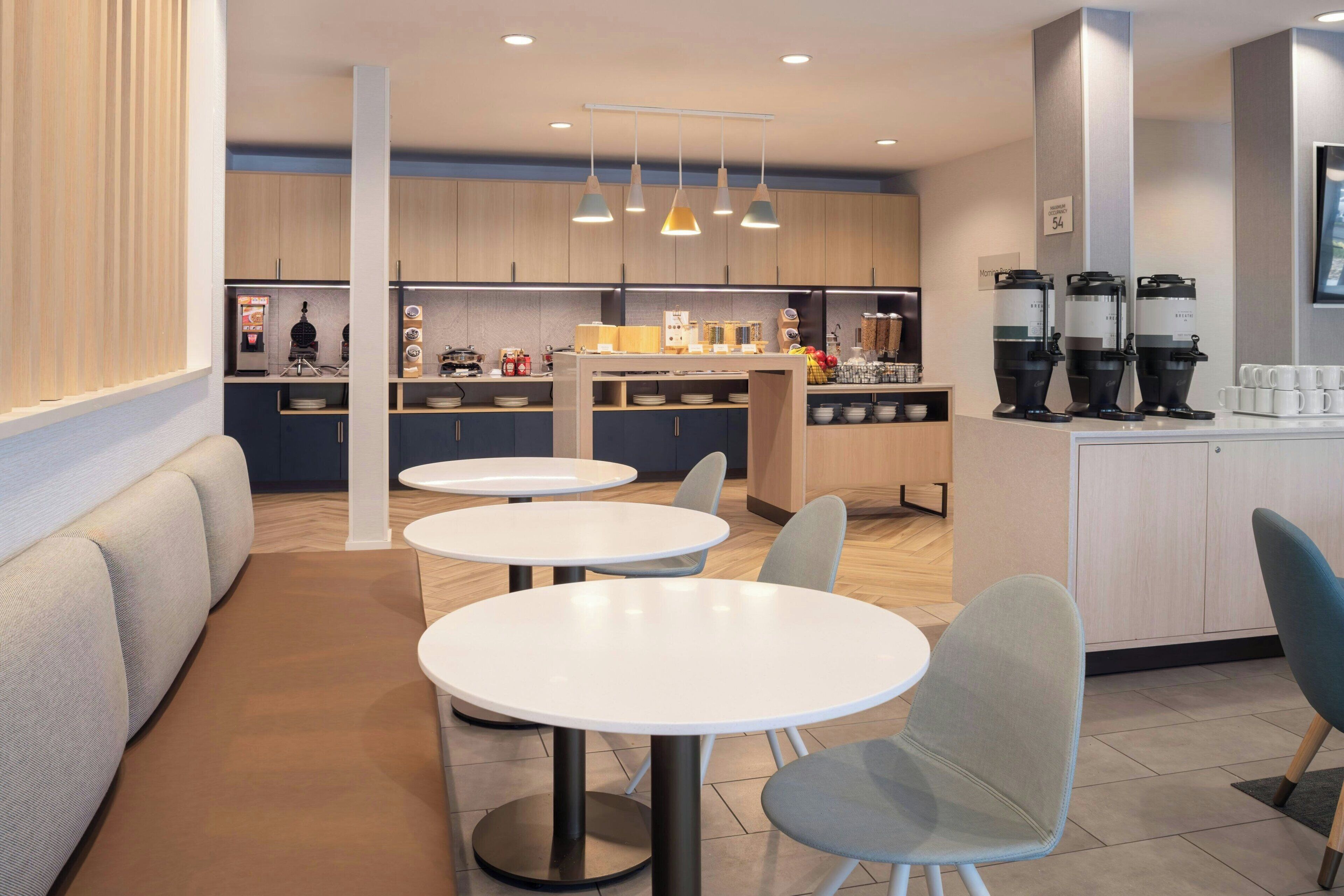 Un café moderne avec des tables et des chaises rondes, un espace cuisine équipé de machines à café et un coin salon avec un canapé.