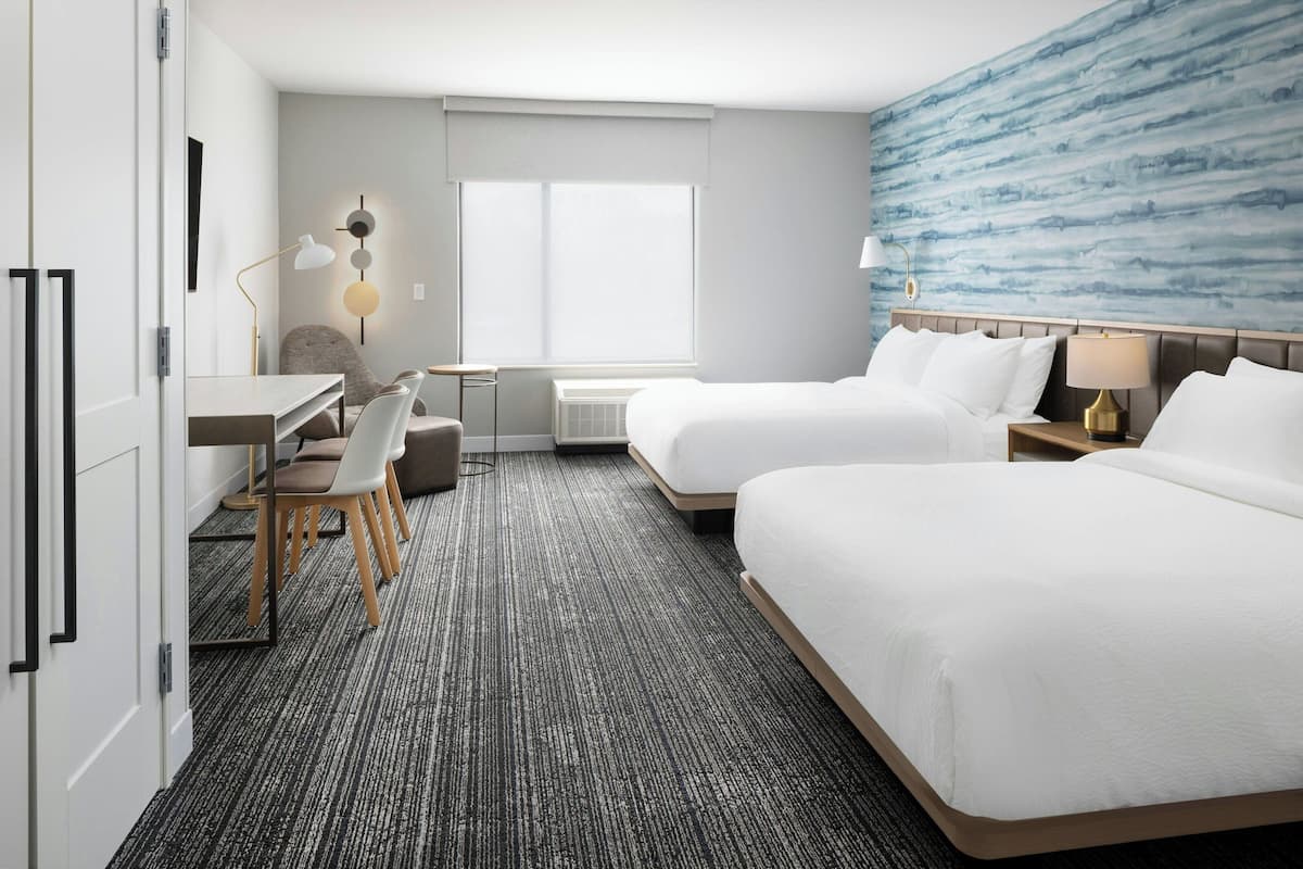 Una habitación de hotel con dos camas, un escritorio, una silla y un ventanal grande.