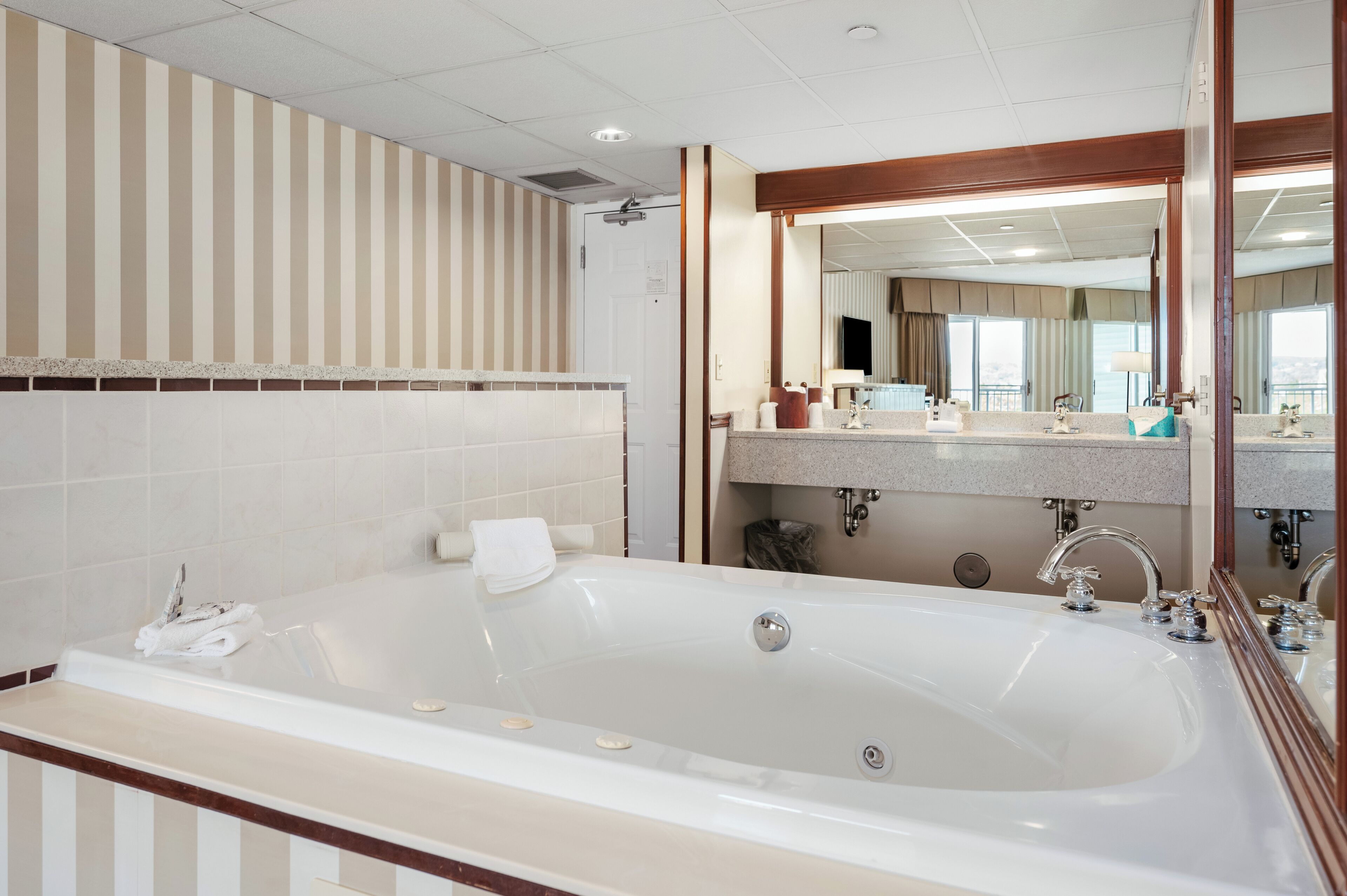 A hotel bathroom with a large bathtub, a vanity with a mirror, and a striped wall design.