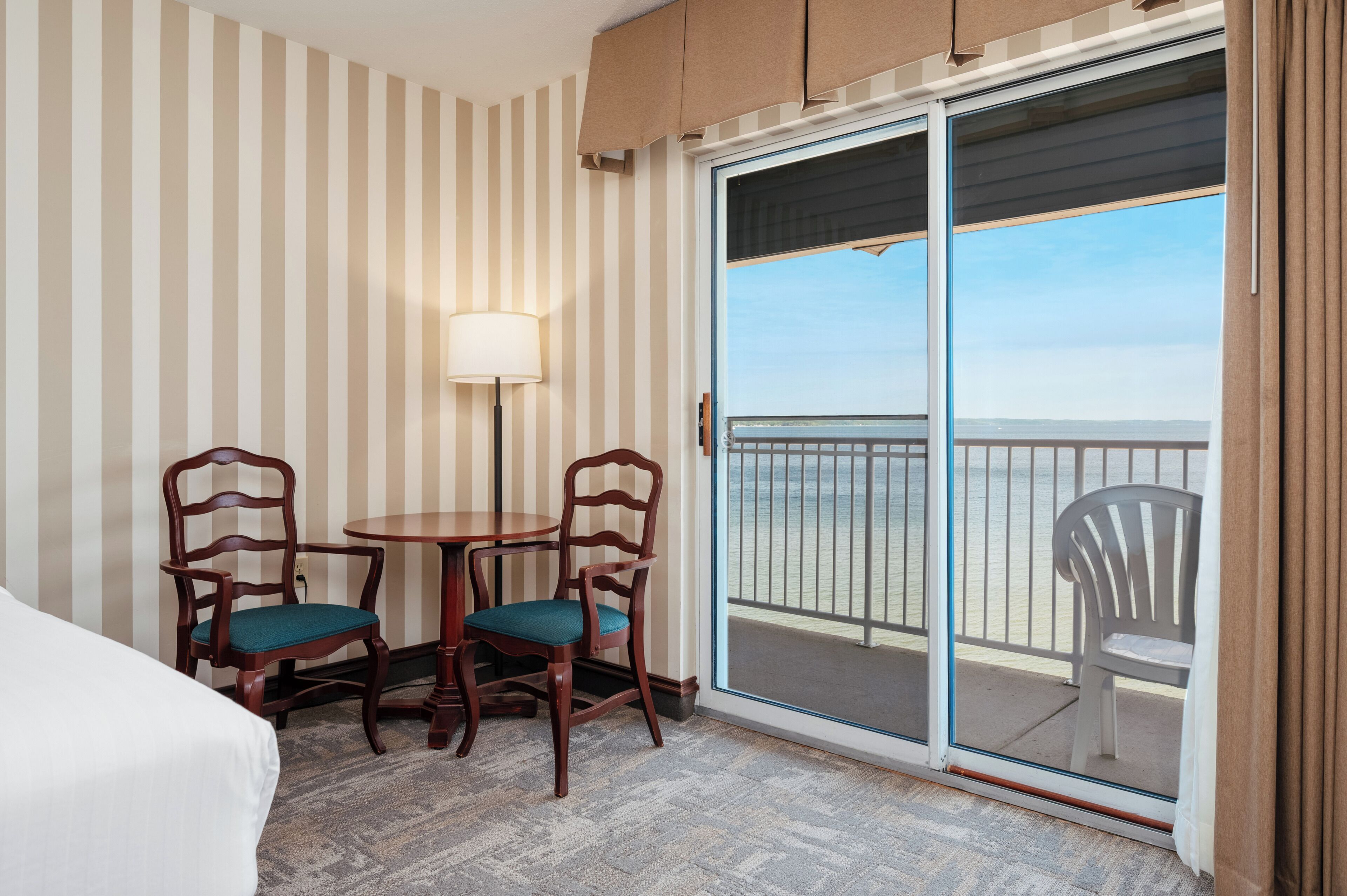 Une chambre d’hôtel avec un papier peint à rayures, un lit, une table avec deux chaises, une lampe et une porte-fenêtre coulissante donnant sur un balcon avec vue sur la plage.