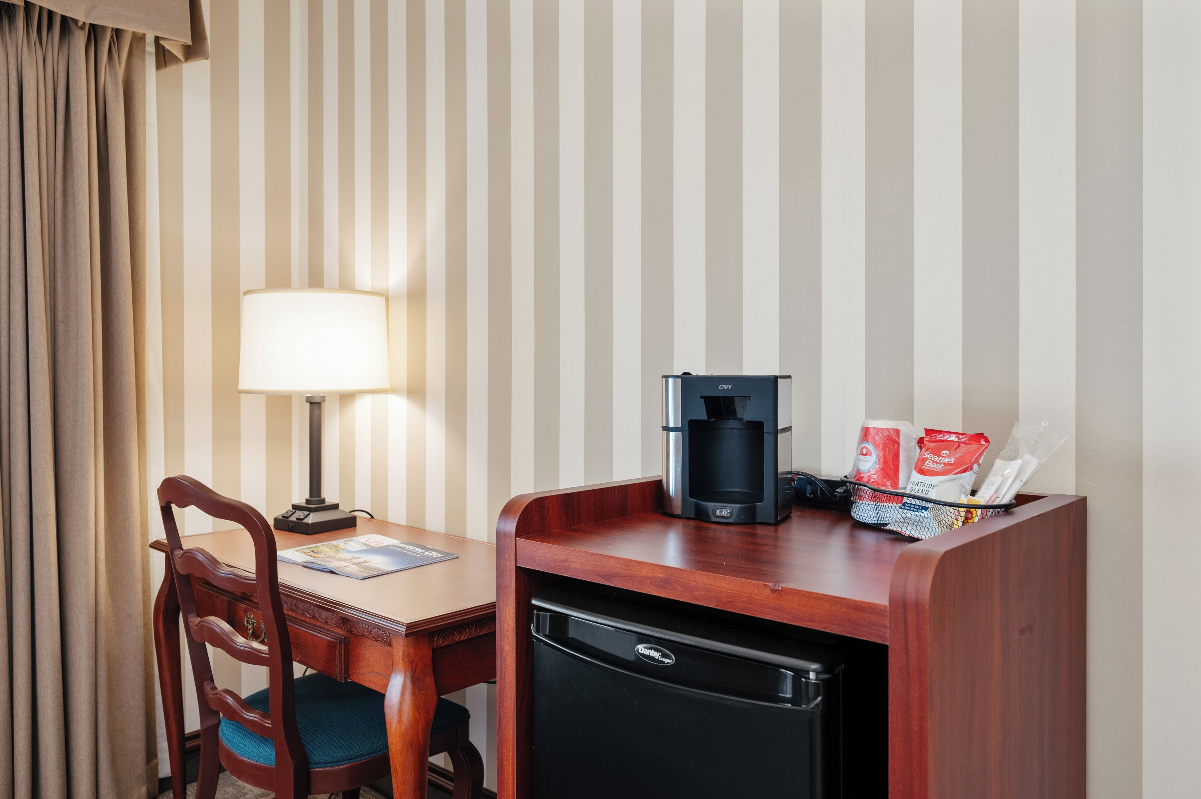 A hotel room with a wooden desk, a lamp, a coffee maker, and a refrigerator.