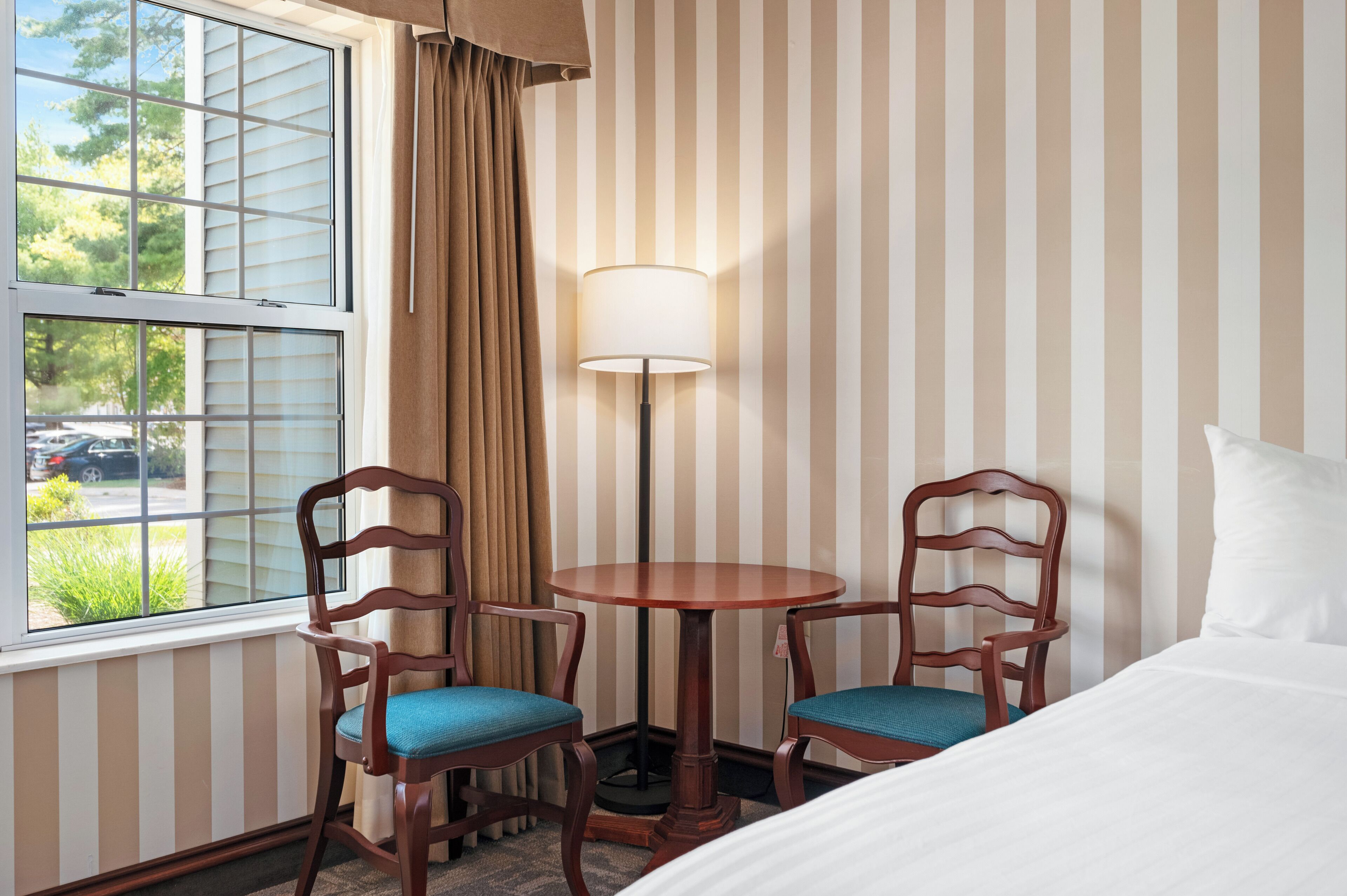 Habitación de hotel con una cama, dos sillas de madera con cojines azules, una mesa redonda, una lámpara de pie, una ventana con persianas y una pared a rayas.