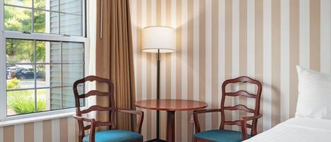 Habitación de hotel con una cama, dos sillas de madera con cojines azules, una mesa redonda, una lámpara de pie, una ventana con persianas y una pared a rayas.