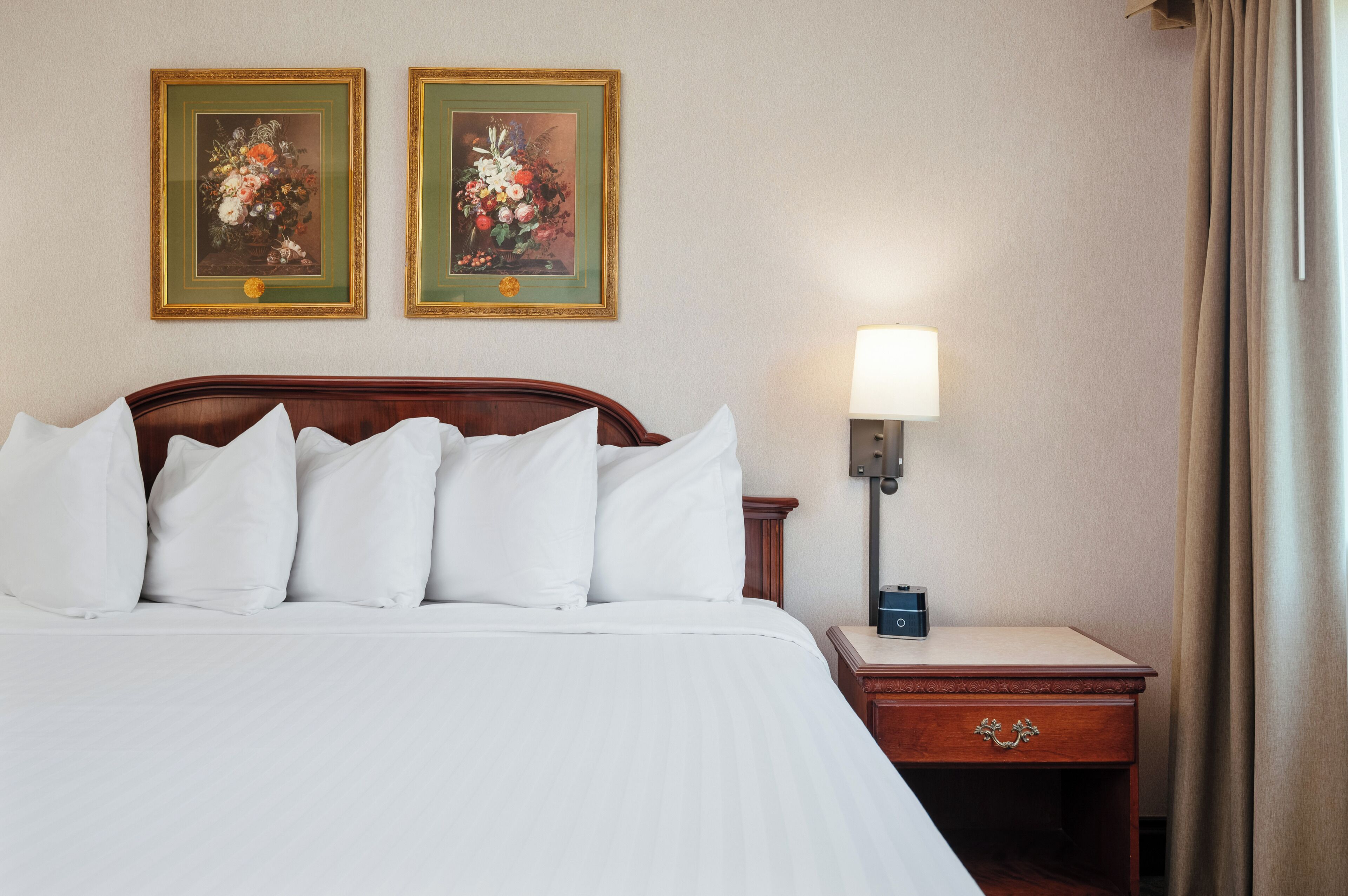 A neatly made bed with white linens, a wooden headboard, a bedside table with a lamp, and two framed floral paintings on the wall.