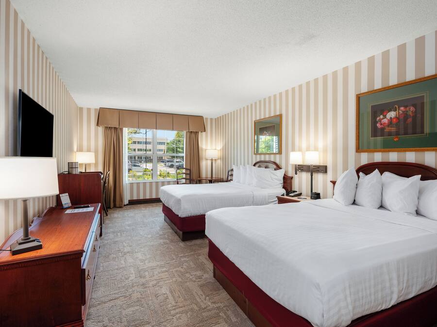 A hotel room with two beds, a desk, a TV, and a window with a view of buildings.