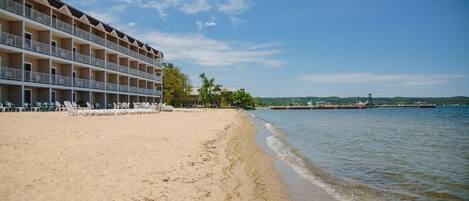 Un hotel frente a la playa con varios balcones, una playa arenosa y vistas al mar y colinas distantes.