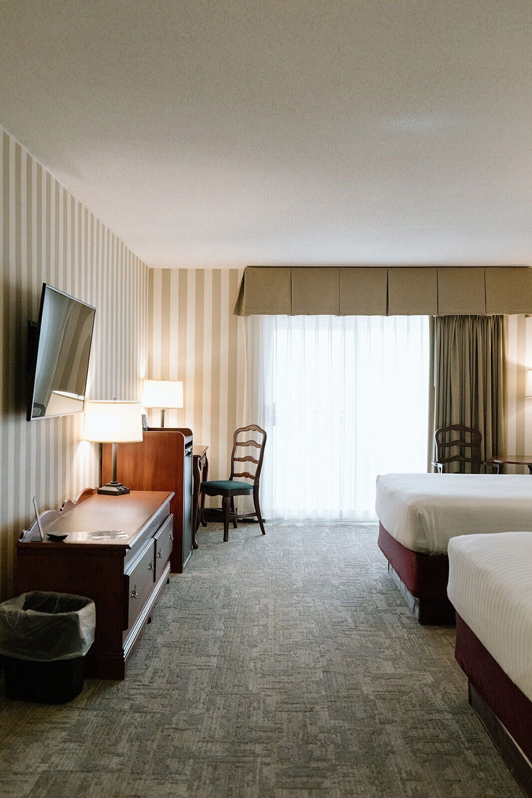 Ein Hotelzimmer mit einem Bett, einem Schreibtisch mit Lampe, einem Sessel, einem Fernseher und einem Fenster mit Vorhängen.