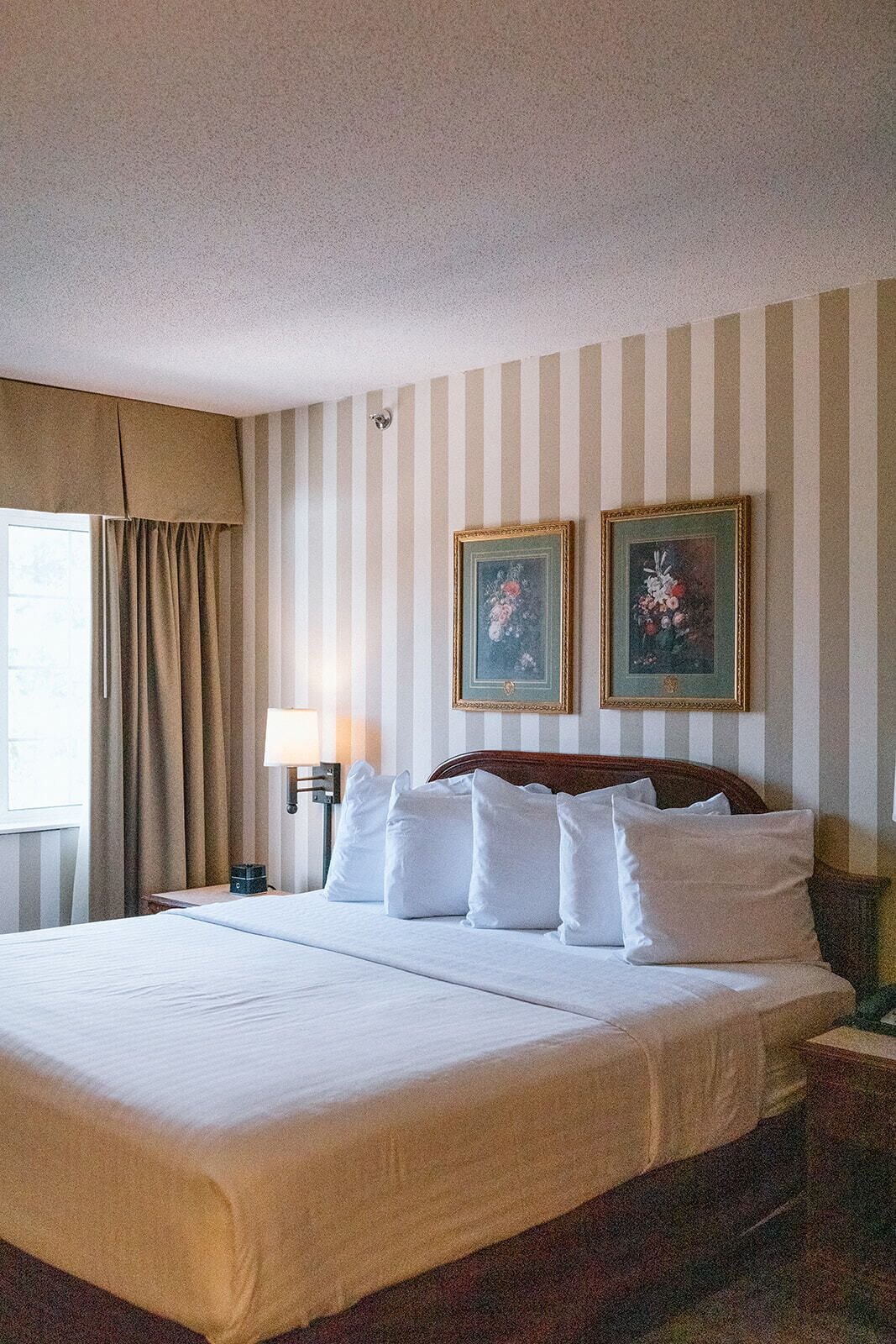 Quarto de hotel com uma cama grande, uma mesa de cabeceira com abajur e dois quadros florais na parede.